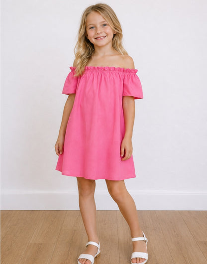 A young girl with blonde hair stands smiling, wearing a bright pink Off-The-Shoulder Short-Sleeve Dress and white sandals, set against a plain white wall and light wood floor.