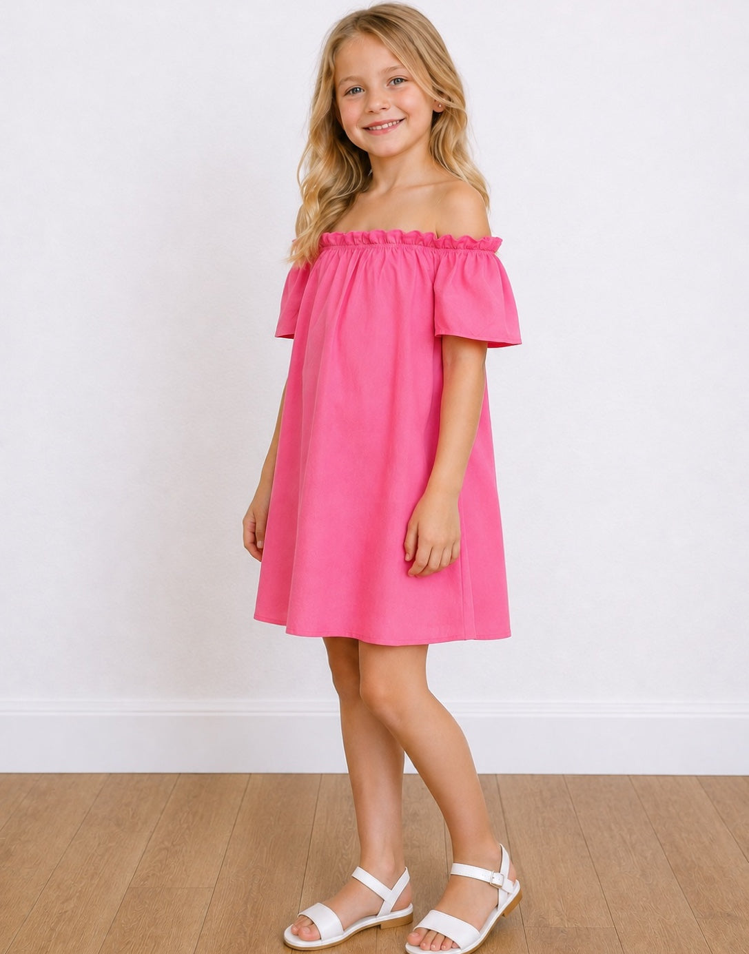 Side view of a young girl with blonde hair stands smiling, wearing a bright pink Off-The-Shoulder Short-Sleeve Dress and white sandals, set against a white wall and light wood floor.
