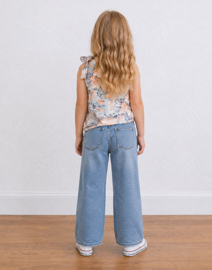Back view of a young girl with blonde hair stands against a white textured wall wearing a sleeveless floral One Shoulder Top with a tie detail, wide-leg blue jeans, and white sneakers.