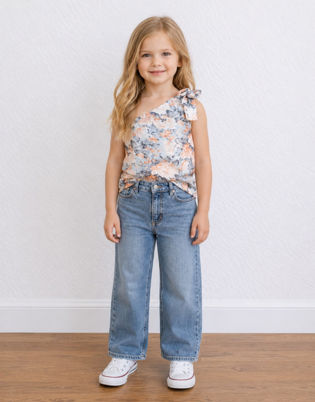 A young girl with blonde hair stands against a white textured wall wearing a sleeveless floral One Shoulder Top with a tie detail, wide-leg blue jeans, and white sneakers.