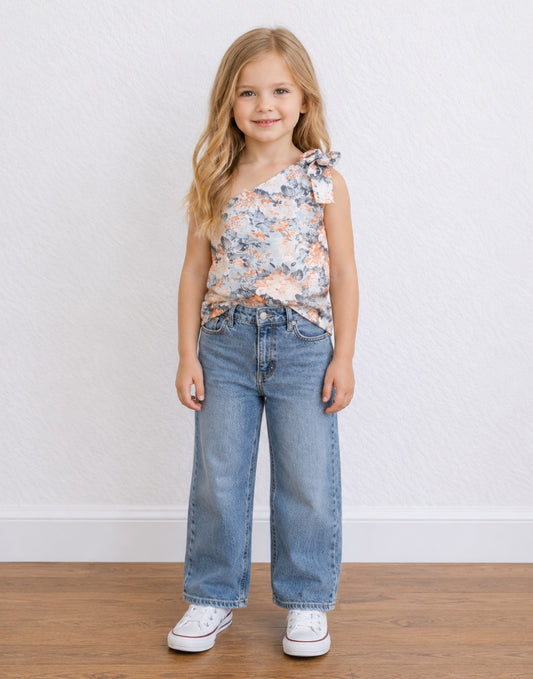 A young girl with blonde hair stands against a white textured wall wearing a sleeveless floral One Shoulder Top with a tie detail, wide-leg blue jeans, and white sneakers.