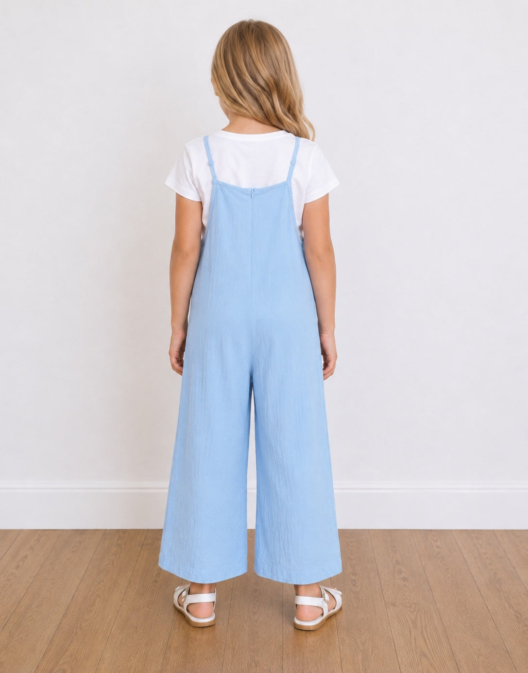 A young girl shown from the back wearing light blue Wide-Leg Overall over a white short-sleeve t-shirt, standing on a light wood floor against a white wall.
