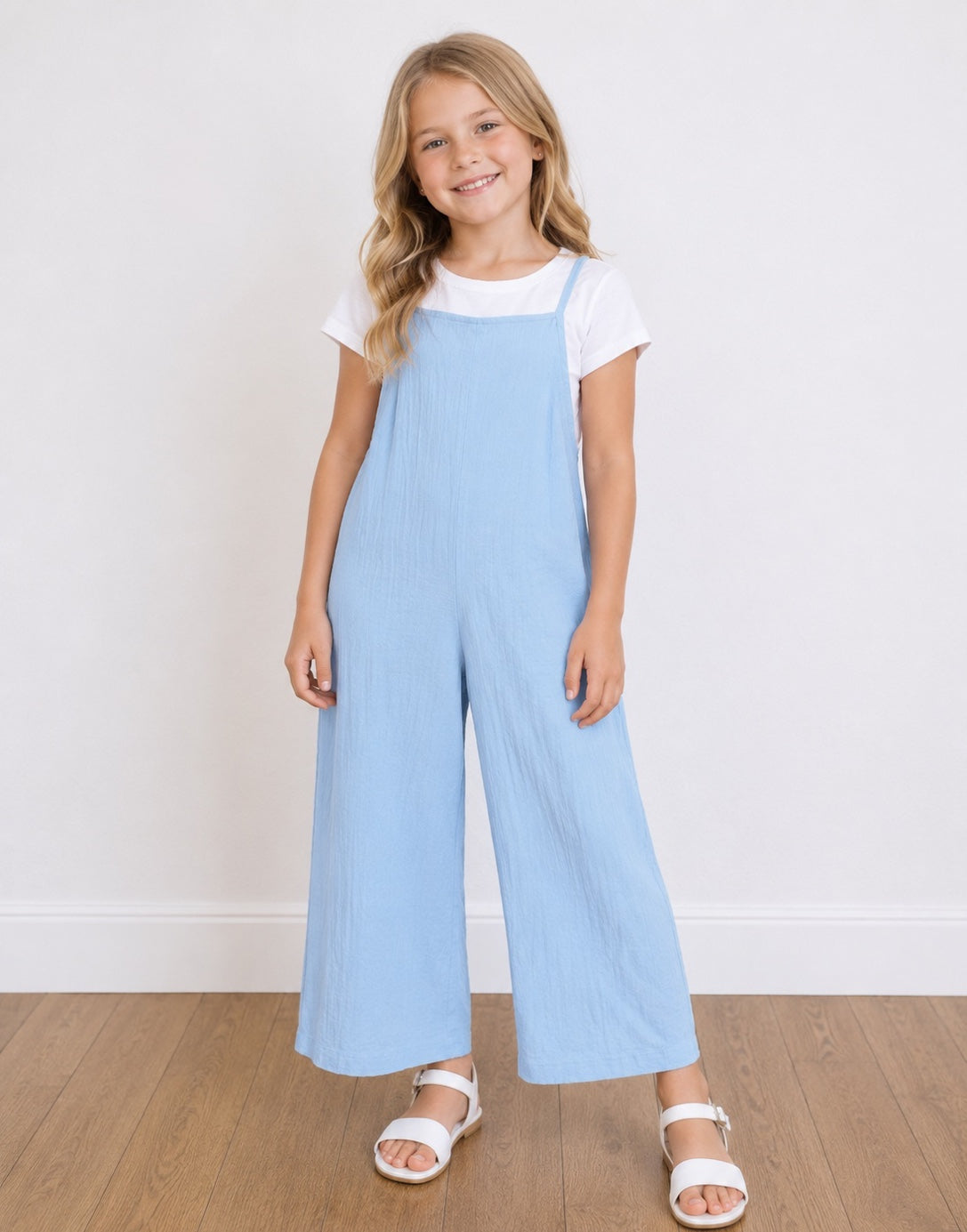 A young girl with blonde hair stands smiling, wearing a light blue Wide-Leg Overall Jumpsuit over a white short-sleeve t-shirt and white sandals, set against a white wall and light wood floor.