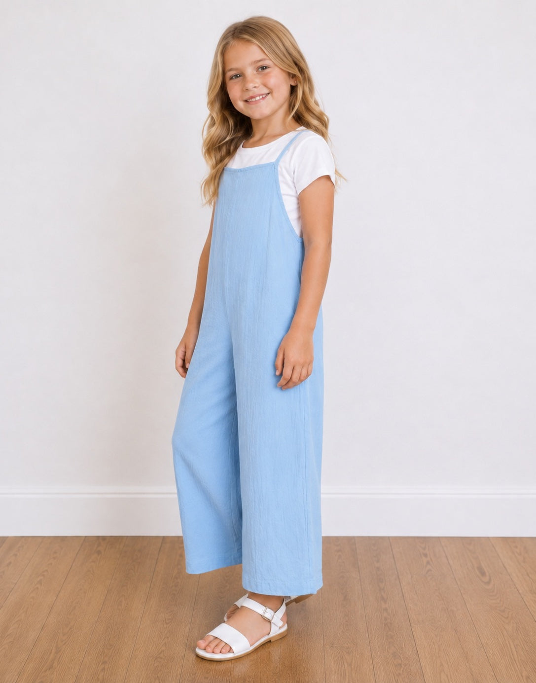 Side view of a young girl with blonde hair stands smiling, wearing a light blue Wide-Leg Jumpsuit over a white short-sleeve t-shirt and white sandals, set against a white wall and light wood floor.