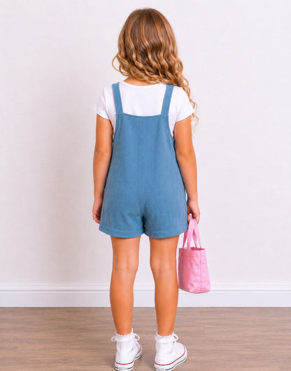 A young girl with wavy light brown hair is shown from behind, wearing a dusty blue short Overall Jumpsuit over a white t-shirt, white sneakers, and holding a pink tote bag against a plain white wall and light wood floor.