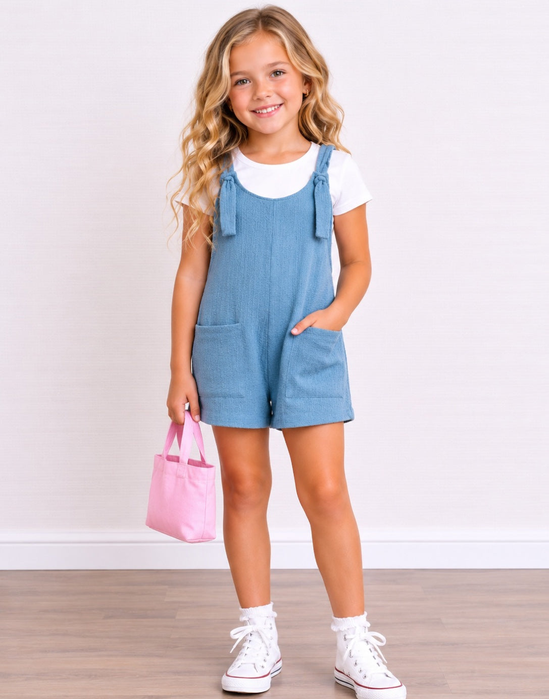 A young girl with blonde wavy hair stands smiling, wearing a light blue denim Overall Romper over a white t-shirt, white socks, and white sneakers. She is holding a small pink tote bag against a white wall and light wood floor background.