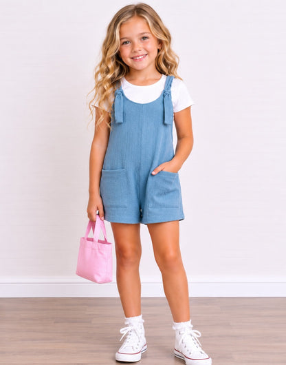 A young girl with blonde wavy hair stands smiling, wearing a light blue denim Overall Romper over a white t-shirt, white socks, and white sneakers. She is holding a small pink tote bag against a white wall and light wood floor background.