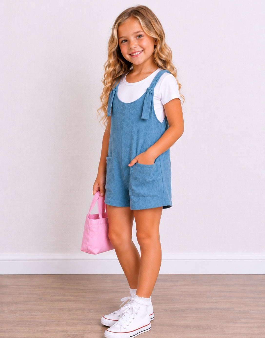 Side view of a young girl with blonde wavy hair stands smiling, wearing a light blue denim Overall Romper over a white t-shirt, white socks, and white sneakers. She is holding a small pink tote bag against a white wall and light wood floor background.
