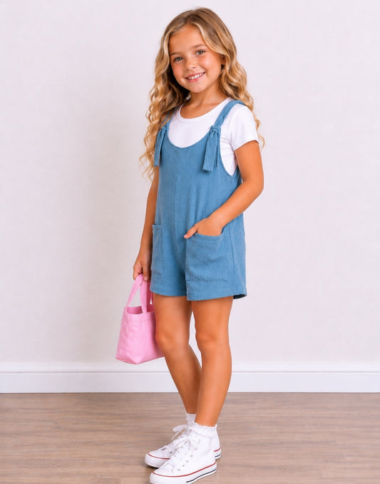 Side view of a young girl with blonde wavy hair stands smiling, wearing a light blue denim Overall Romper over a white t-shirt, white socks, and white sneakers. She is holding a small pink tote bag against a white wall and light wood floor background.