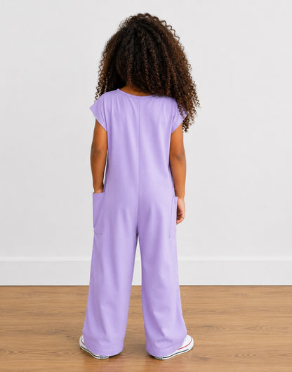Back view of young girl wearing a purple Oversized Knit Tee Jumpsuit with white shoes on a wooden floor with plain background.