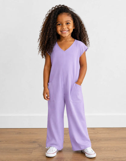 Young girl wearing a purple Oversized Knit Tee Jumpsuit with white shoes on a wooden floor with plain background.