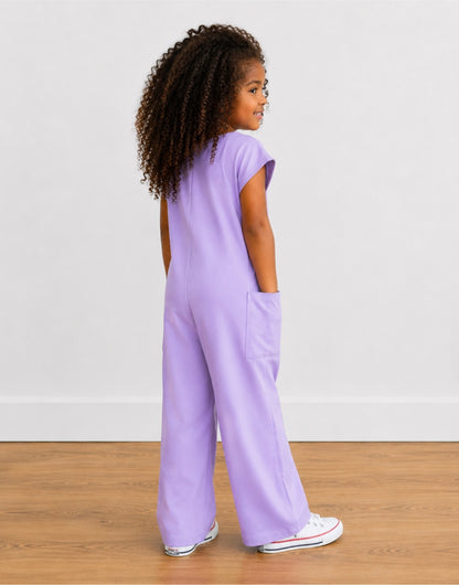 Side view of young girl wearing a purple Oversized Knit Tee Jumpsuit with white shoes on a wooden floor with plain background.