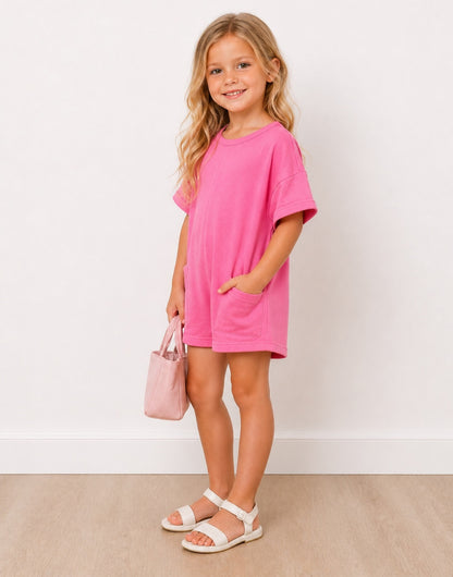 Side view of young girl wearing a pink Oversized Tee Shirt Romper with a matching handbag against a white background.