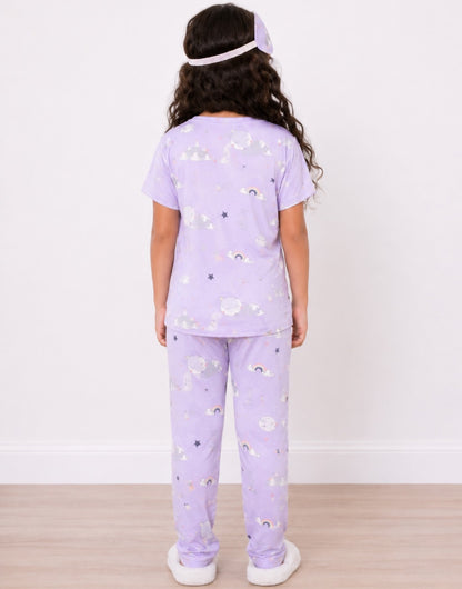 Back view of a young girl stands against a white wall wearing a light purple Pajama Set with a moon and star print, a matching sleep mask, and white slippers.