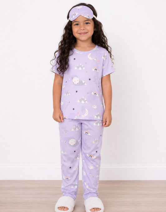 A young girl stands against a white wall wearing a light purple Pajama Set with a moon and star print, a matching sleep mask, and white slippers.