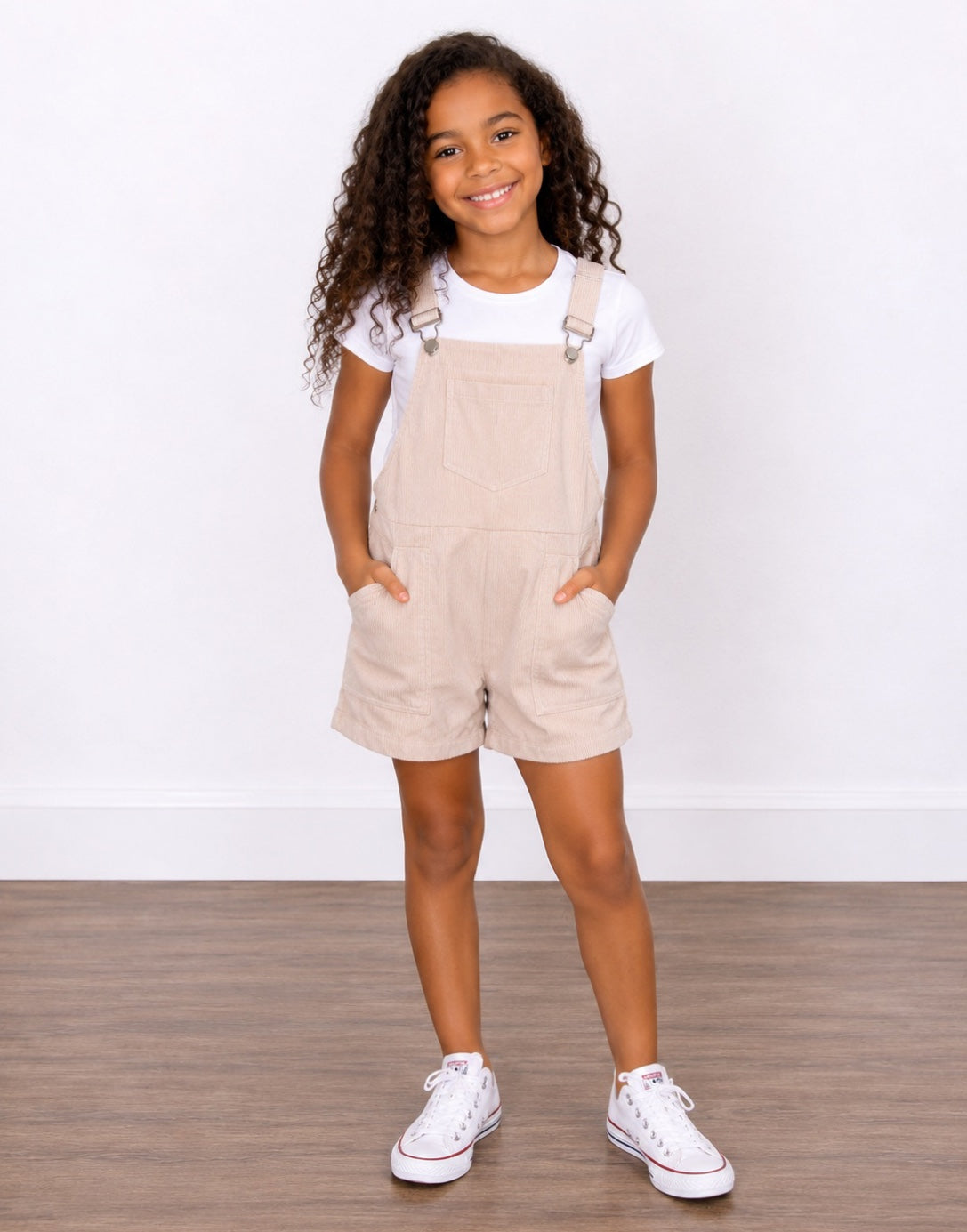 A young girl with curly brown hair wearing light beige corduroy Pocket Overall Romper over a white t-shirt, paired with white sneakers. She is posing with her hands in the pockets of the overalls against a white wall and wood floor background.