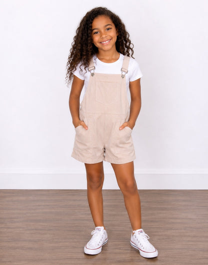 A young girl with curly brown hair wearing light beige corduroy Pocket Overall Romper over a white t-shirt, paired with white sneakers. She is posing with her hands in the pockets of the overalls against a white wall and wood floor background.