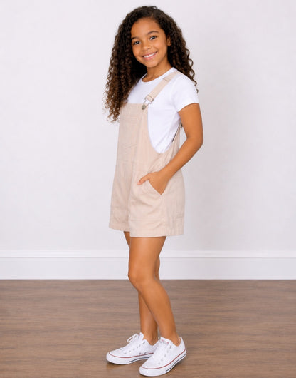 Side view of a young girl with curly brown hair wearing light beige corduroy Pocket Overall Romper over a white t-shirt, paired with white sneakers. She is posing with her hands in the pockets of the overalls against a white wall and wood floor background.