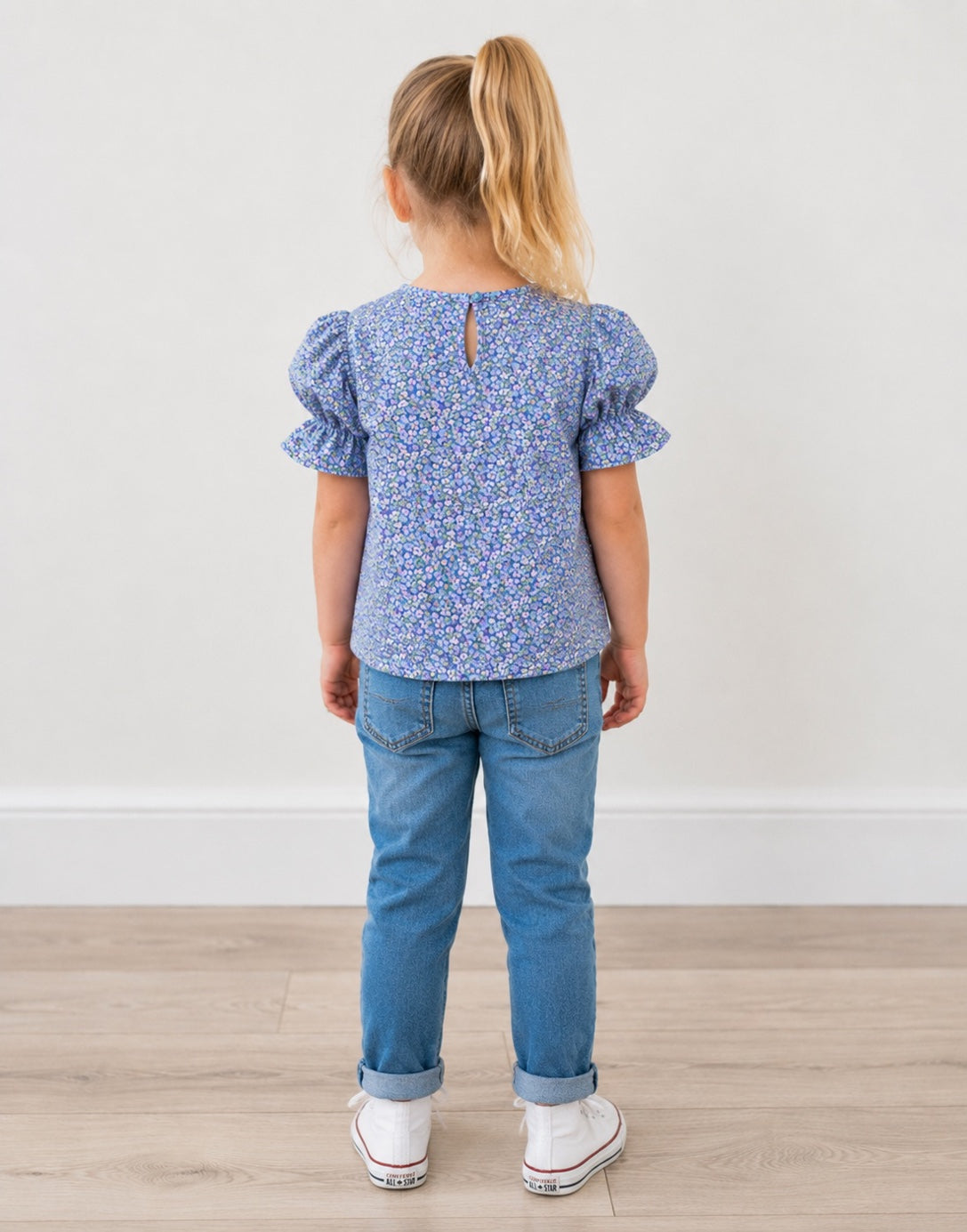 Back view of a young girl with blonde hair in a ponytail is shown from behind, wearing a blue and white floral patterned puff-sleeve top, blue jeans, and white sneakers, standing against a plain white wall and light wood floor.