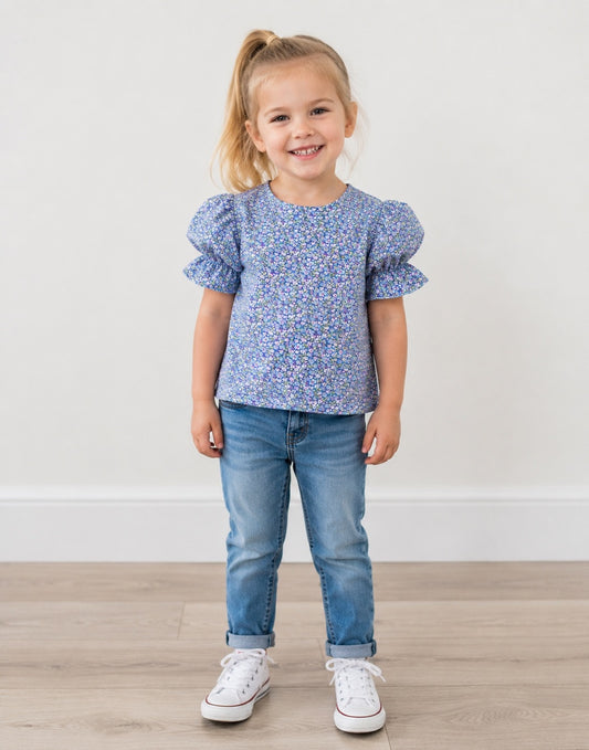 A young girl with blonde hair in a ponytail stands smiling, wearing a blue and white floral print Puff Sleeve Blouse, blue jeans, and white sneakers against a plain white wall and light wood floor.
