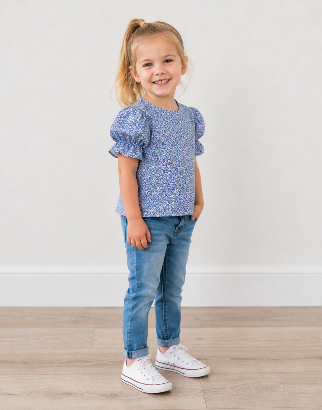 Side view of a young girl with blonde hair in a ponytail stands smiling, wearing a blue and white floral puff-sleeve top, blue jeans, and white sneakers against a neutral indoor background.