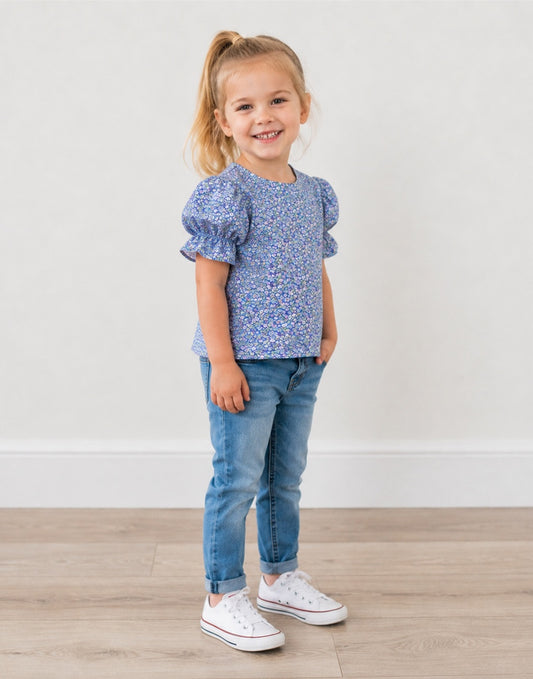 Side view of a young girl with blonde hair in a ponytail stands smiling, wearing a blue and white floral puff-sleeve top, blue jeans, and white sneakers against a neutral indoor background.