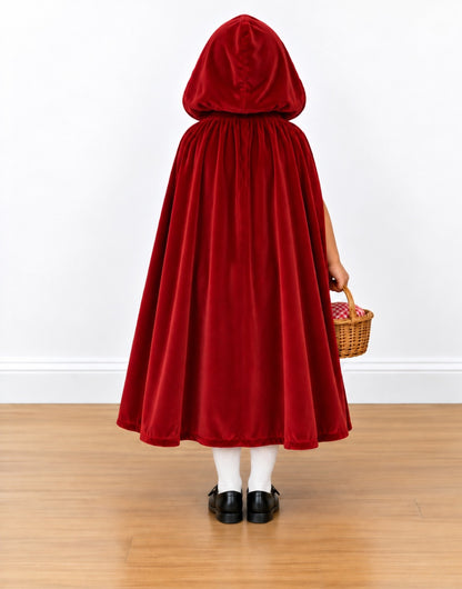 A child wearing a red velvet hooded cape, shown from the back, holding a small woven basket against a white wall and light wood floor.