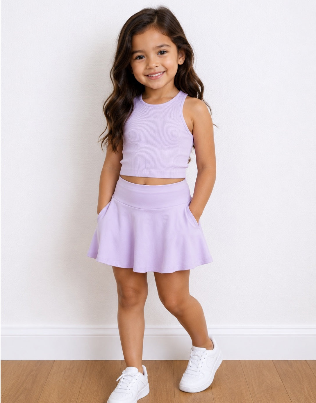 A young girl with long brown hair stands in a studio wearing a light purple/lavender sleeveless crop top and matching flared Sport Skort, paired with white sneakers, against a white wall and wood floor.