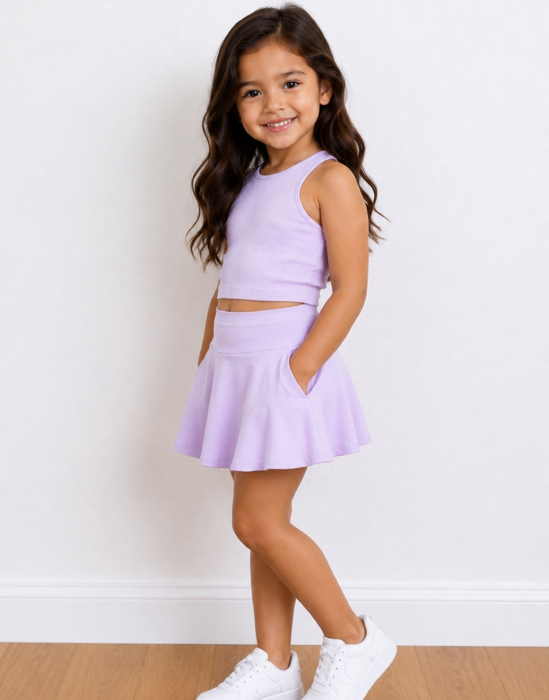 Side view of a young girl with long brown hair stands in a studio wearing a light purple/lavender two-piece outfit consisting of a sleeveless crop top and a flared Sport Skort with side pockets, paired with white sneakers.