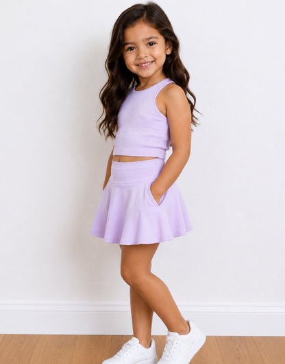 Side view of a young girl with long brown hair stands in a studio wearing a light purple/lavender two-piece outfit consisting of a sleeveless crop top and a flared Sport Skort with side pockets, paired with white sneakers.