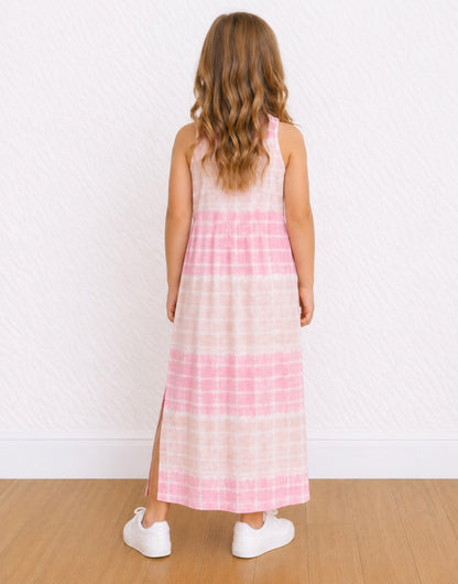 A young girl with long wavy brown hair is shown from behind wearing a sleeveless pink and white tie-dye Maxi Tank Dress with a side slit, paired with white sneakers, standing against a textured white wall and light wood floor.