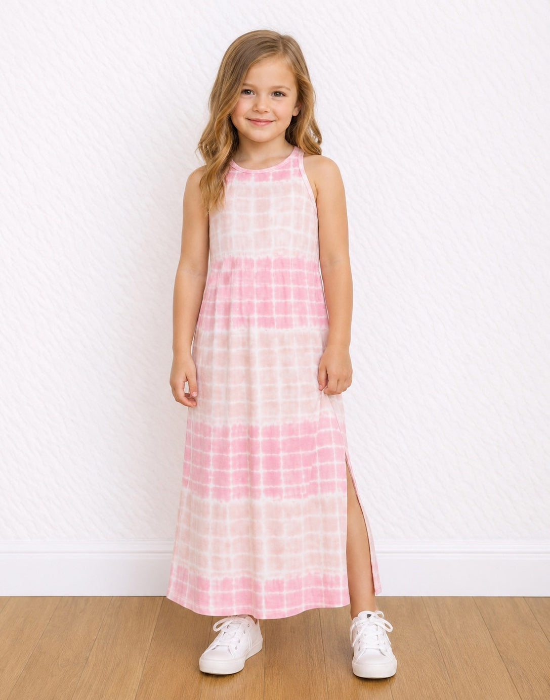 A young girl stands on a light wood floor against a white textured wall, wearing a sleeveless pink and white tie-dye Maxi Tank Dress with a high neckline and side slit, paired with white sneakers.