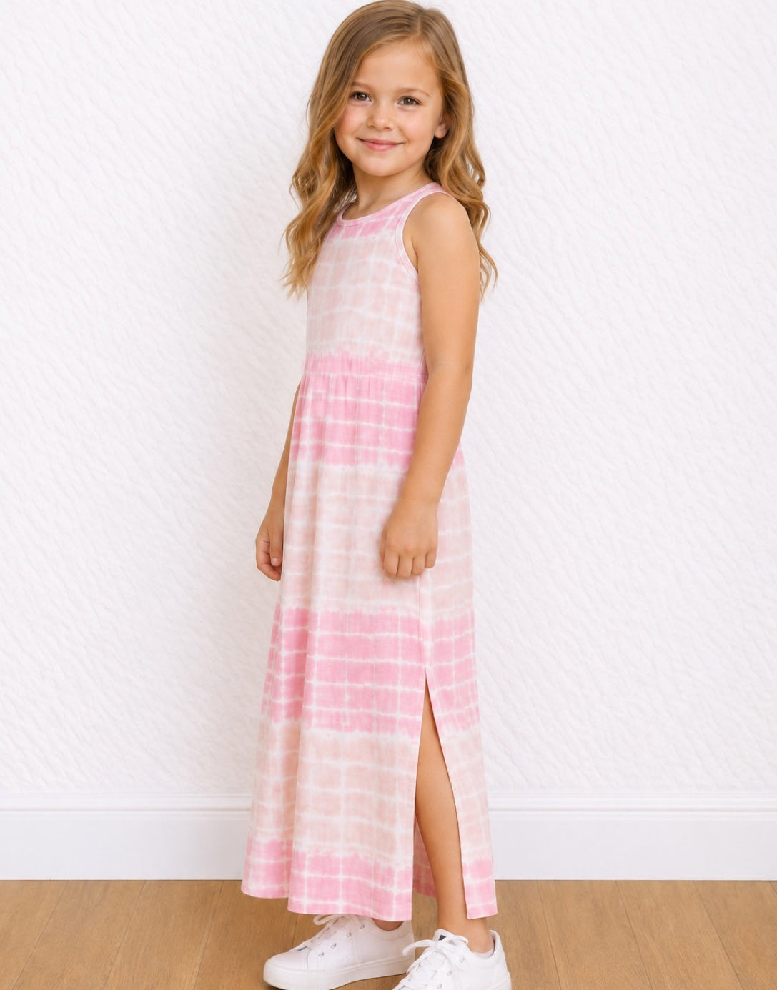 A young girl with blonde hair stands in a studio wearing a sleeveless pink and white tie-dye Maxi Tank Dress with a side slit, paired with white sneakers.