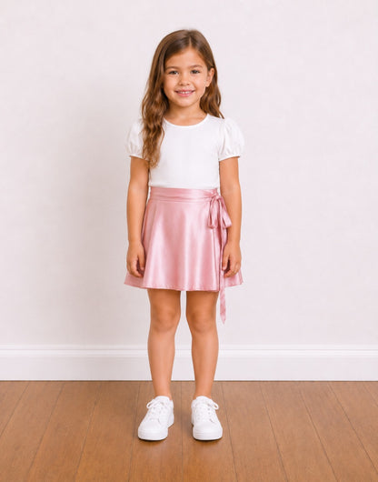 A young girl stands on a wooden floor against a white wall, wearing a white short-sleeve top and a shiny light pink satin Wrap Mini Skirt with a side tie.