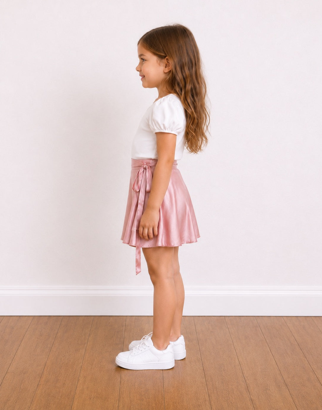 Side view of a young girl stands on a wooden floor against a white wall, wearing a white short-sleeve top and a shiny light pink satin Wrap Mini Skirt with a side tie.