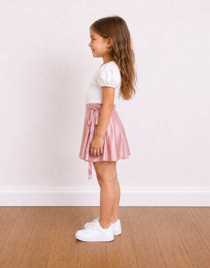 Side view of a young girl stands on a wooden floor against a white wall, wearing a white short-sleeve top and a shiny light pink satin Wrap Mini Skirt with a side tie.