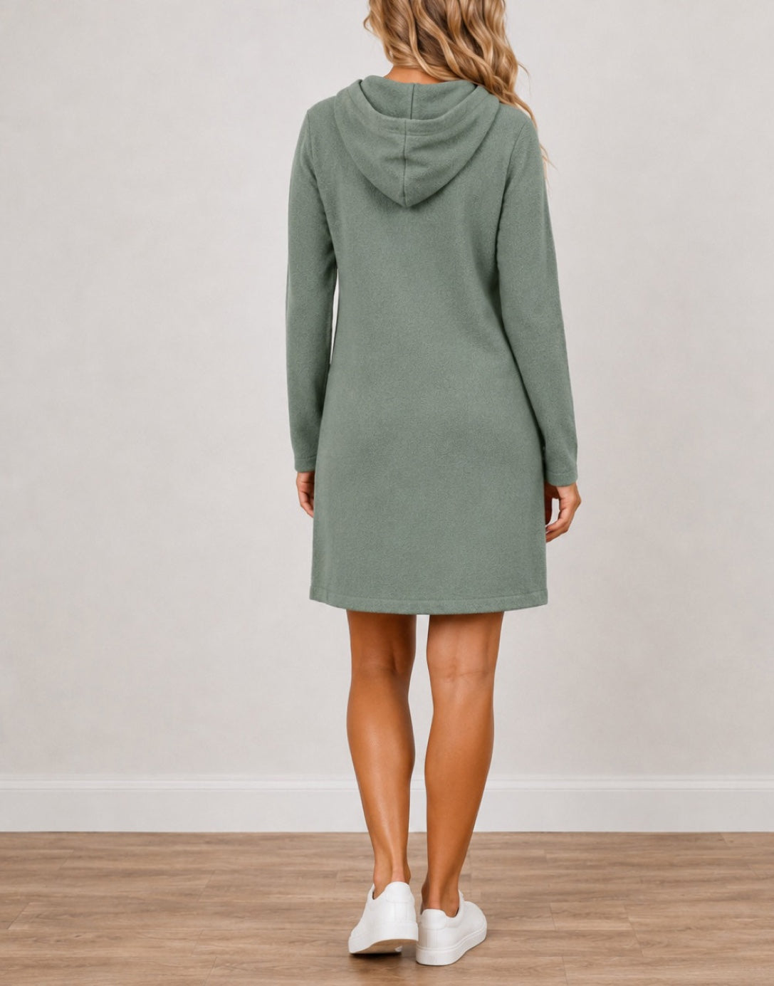 A woman viewed from the back wearing a sage green Hooded Long-Sleeve Mini Dress, paired with white sneakers, standing on a light wood floor against a neutral off-white wall.