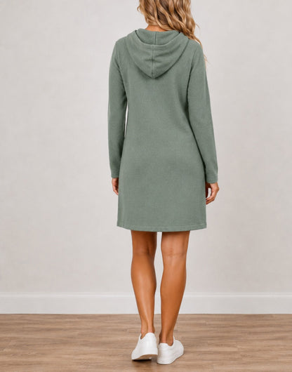 A woman viewed from the back wearing a sage green Hooded Long-Sleeve Mini Dress, paired with white sneakers, standing on a light wood floor against a neutral off-white wall.