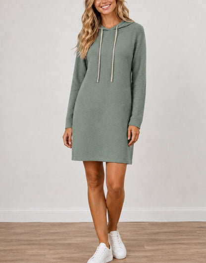 A woman wearing a sage green Long-Sleeve Knit Hoodie Mini Dress with beige drawstrings, paired with white sneakers, standing against a neutral light gray wall and wood floor.