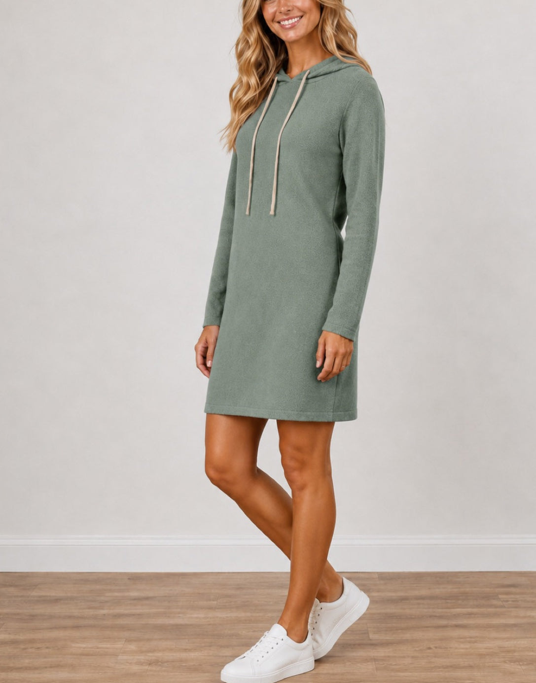 Side view of a woman wearing a sage green Long-Sleeve Hooded Knit Mini Dress with beige drawstrings, paired with white sneakers, standing against a neutral studio background.