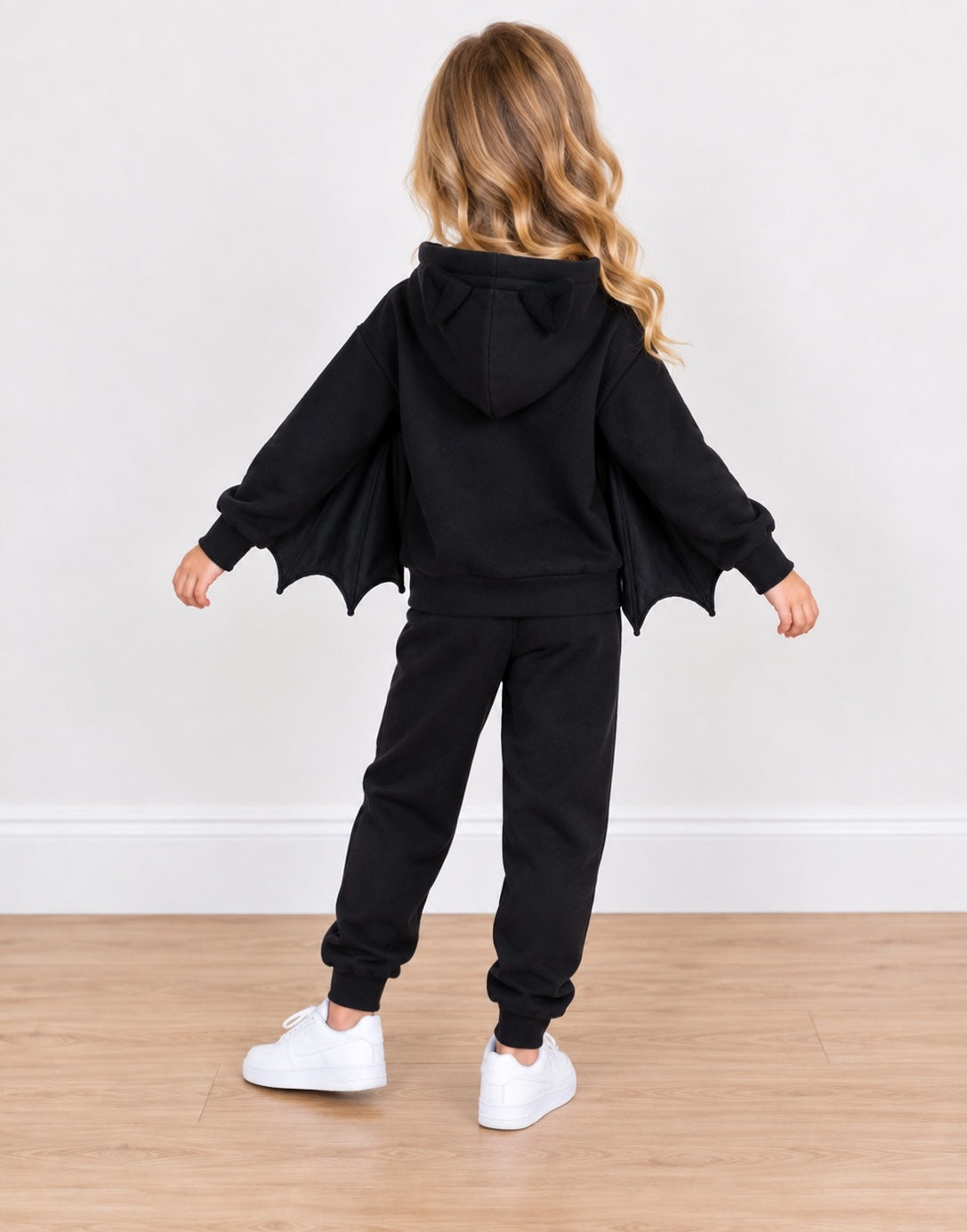 Back view of a young girl wearing a black bat-themed zip-up hoodie and matching sweatpants set, featuring a hood and attached batwings, paired with white sneakers against a plain indoor wall and wood floor.