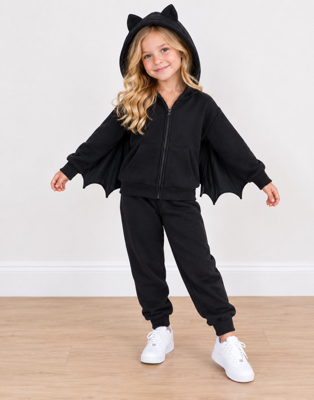 A young girl wearing a black bat-themed zip-up hoodie and matching sweatpants set, featuring a hood with cat ears and attached bat wings, paired with white sneakers against a plain indoor wall and wood floor.