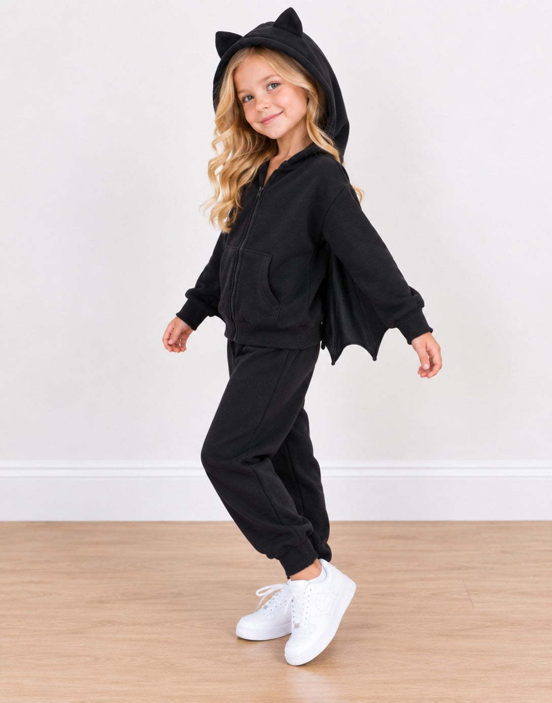Side view of a young girl wearing a black batwing zip-up hoodie and matching sweatpants set, featuring a hood with cat ears and attached bat wings, paired with white sneakers against a plain indoor wall and wood floor.