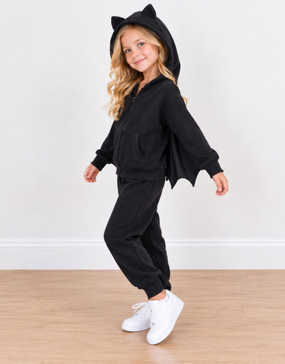 Side view of a young girl wearing a black batwing zip-up hoodie and matching sweatpants set, featuring a hood with cat ears and attached bat wings, paired with white sneakers against a plain indoor wall and wood floor.