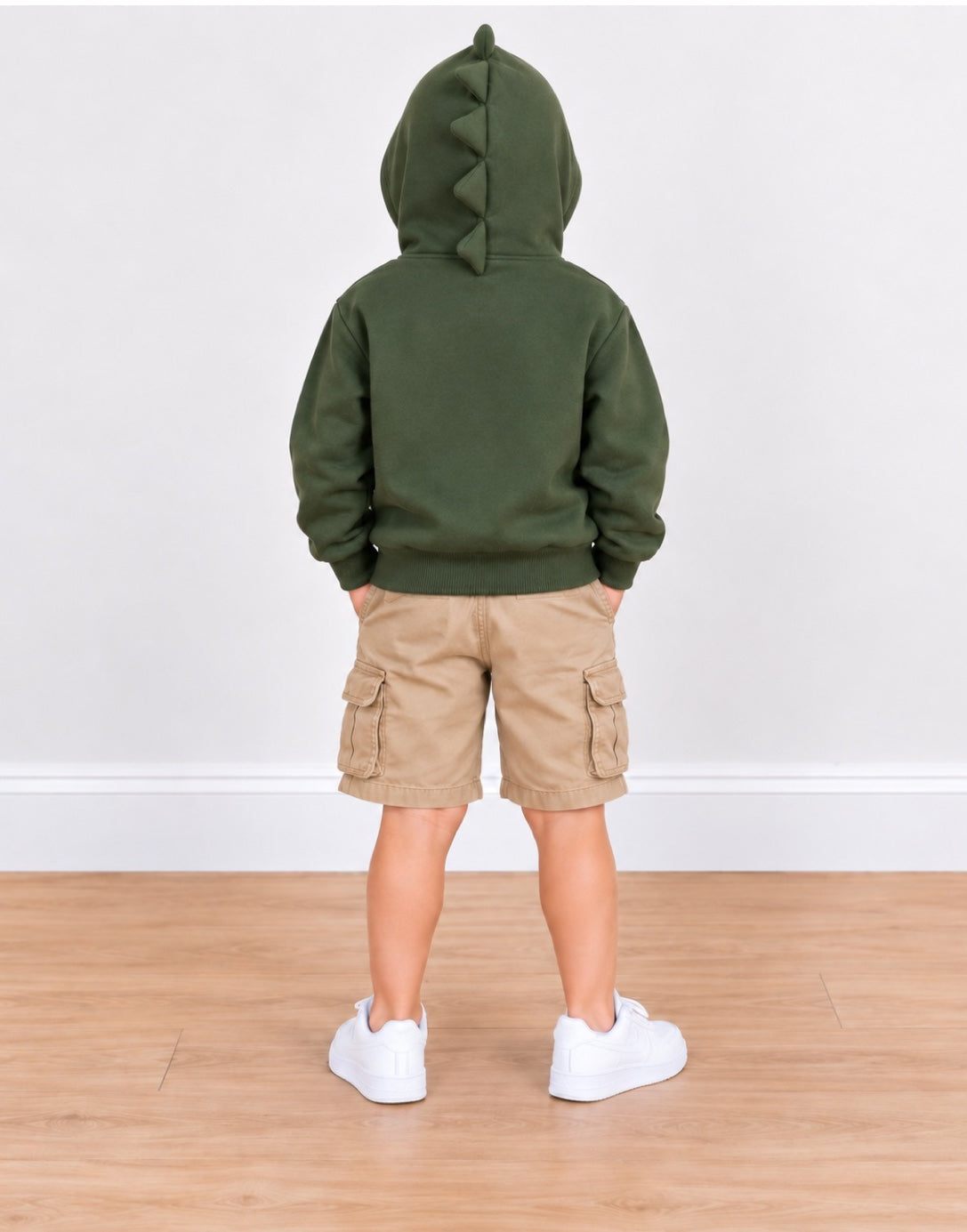 A child standing with their back to the camera, wearing an olive green hooded sweatshirt with a dinosaur-style tail detail on the hood, tan cargo shorts, and white sneakers, set against a plain white wall and light wood floor.