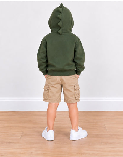 A child standing with their back to the camera, wearing an olive green hooded sweatshirt with a dinosaur-style tail detail on the hood, tan cargo shorts, and white sneakers, set against a plain white wall and light wood floor.
