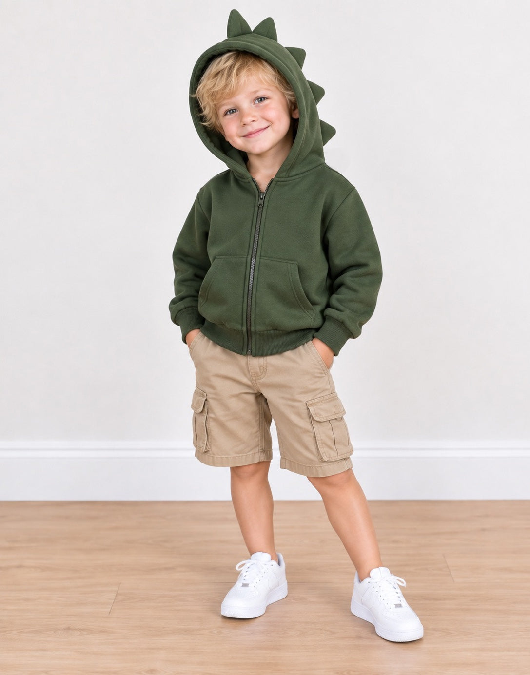 A young child wearing an olive green zip-up hoodie with a dinosaur-style hood and shoulder spikes, paired with khaki cargo shorts and white sneakers, standing against a plain white wall and light wood floor.