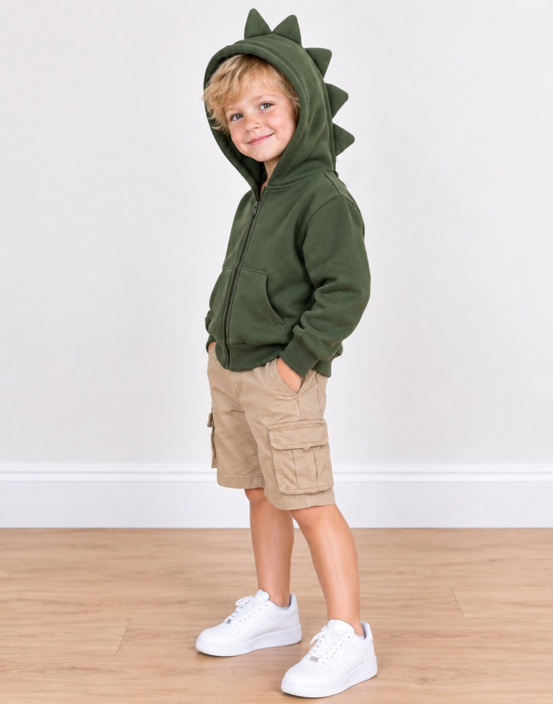 Side view of a young child wearing an olive green zip-up hoodie with dinosaur-style spikes on the hood, paired with khaki cargo shorts and white sneakers, standing against a plain white wall and light wood floor.