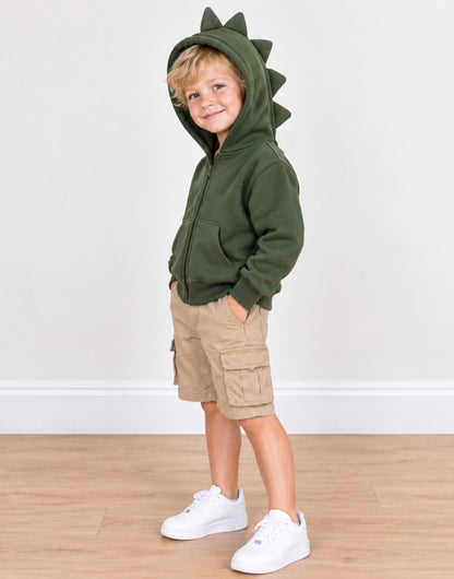 Side view of a young child wearing an olive green zip-up hoodie with dinosaur-style spikes on the hood, paired with khaki cargo shorts and white sneakers, standing against a plain white wall and light wood floor.