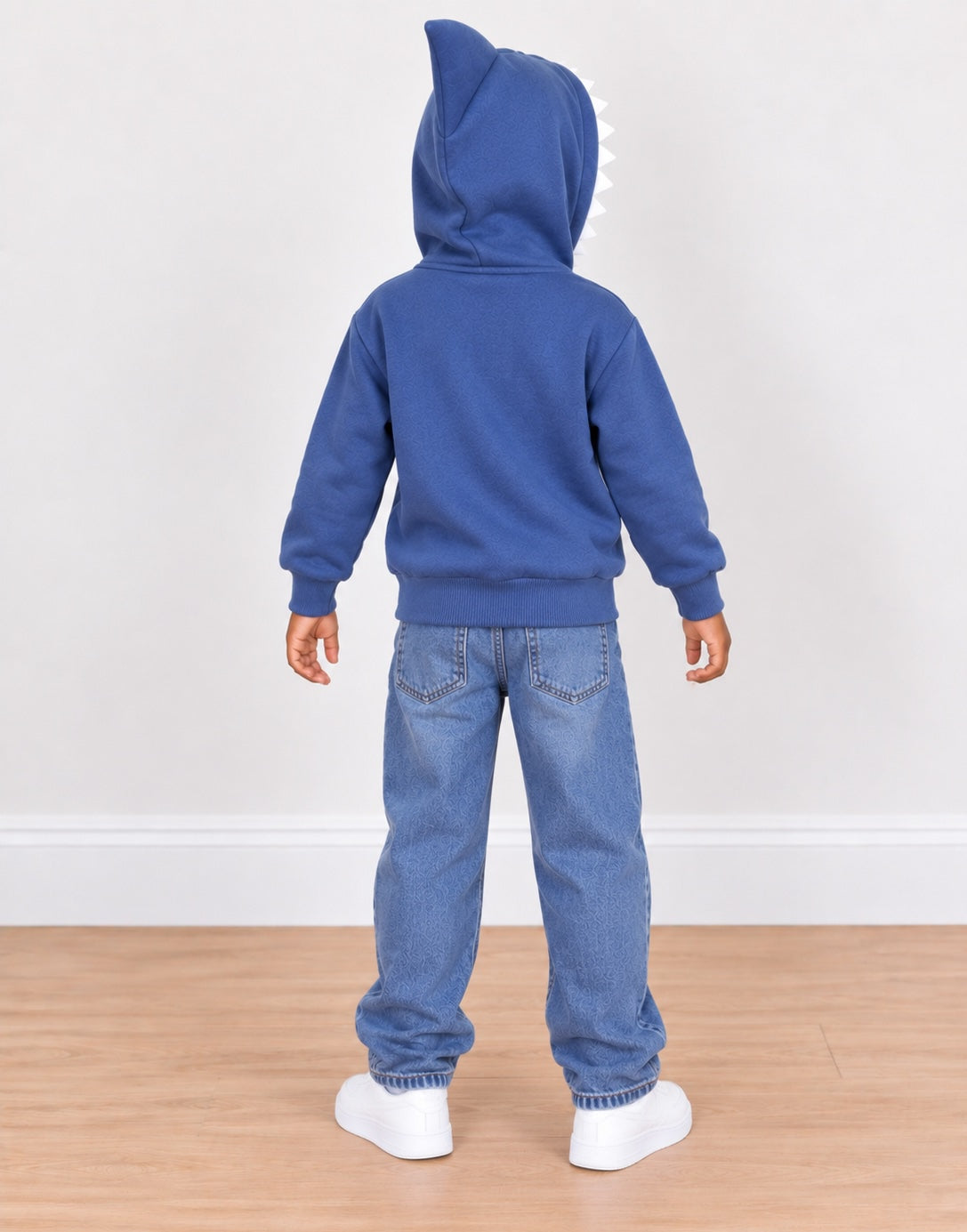 A child standing with their back to the camera, wearing a royal blue hooded sweatshirt with a shark-fin detail on the hood, blue jeans, and white sneakers, set against a plain white wall and light wood floor.